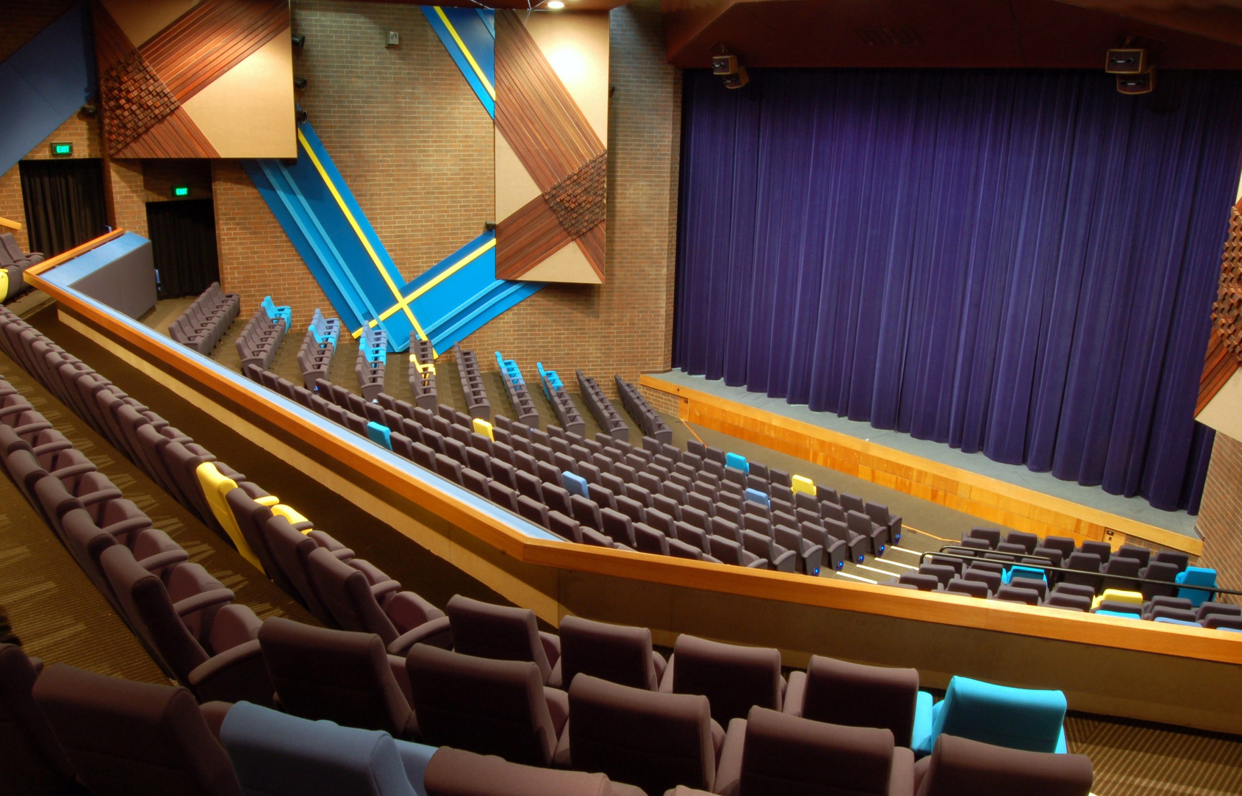 The interior of the Chaffey Theatre, shot from the audience's perspective. The theatre is empty and dark, with brown seats and a deep blue curtain closed across the stage area.