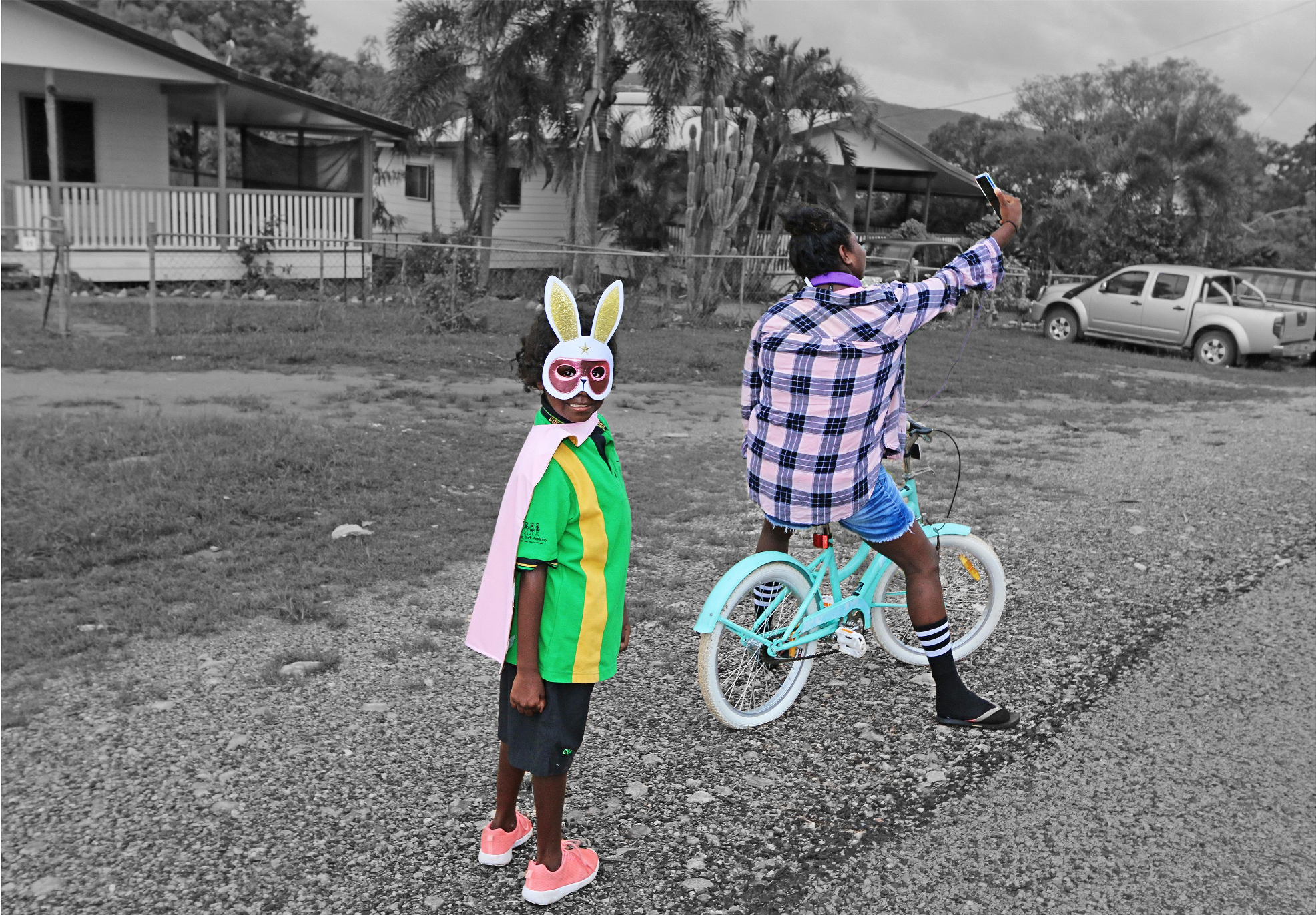 A young Indigenous girl in a pink and purple flannelette shirt is sitting on a bike facing away from the camera, taking a selfie. A younger Indigenous kid is looking at the camera, wearing a pink cape and a glittery rabbit mask.