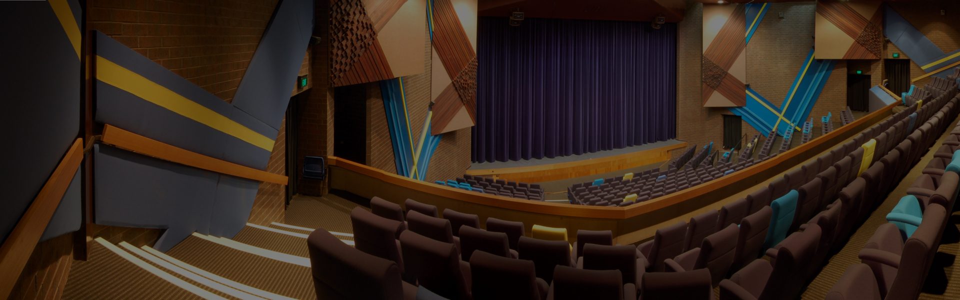 The interior of the Chaffey Theatre, shot from the audience's perspective. The theatre is empty and dark, with brown seats and a deep blue curtain closed across the stage area.